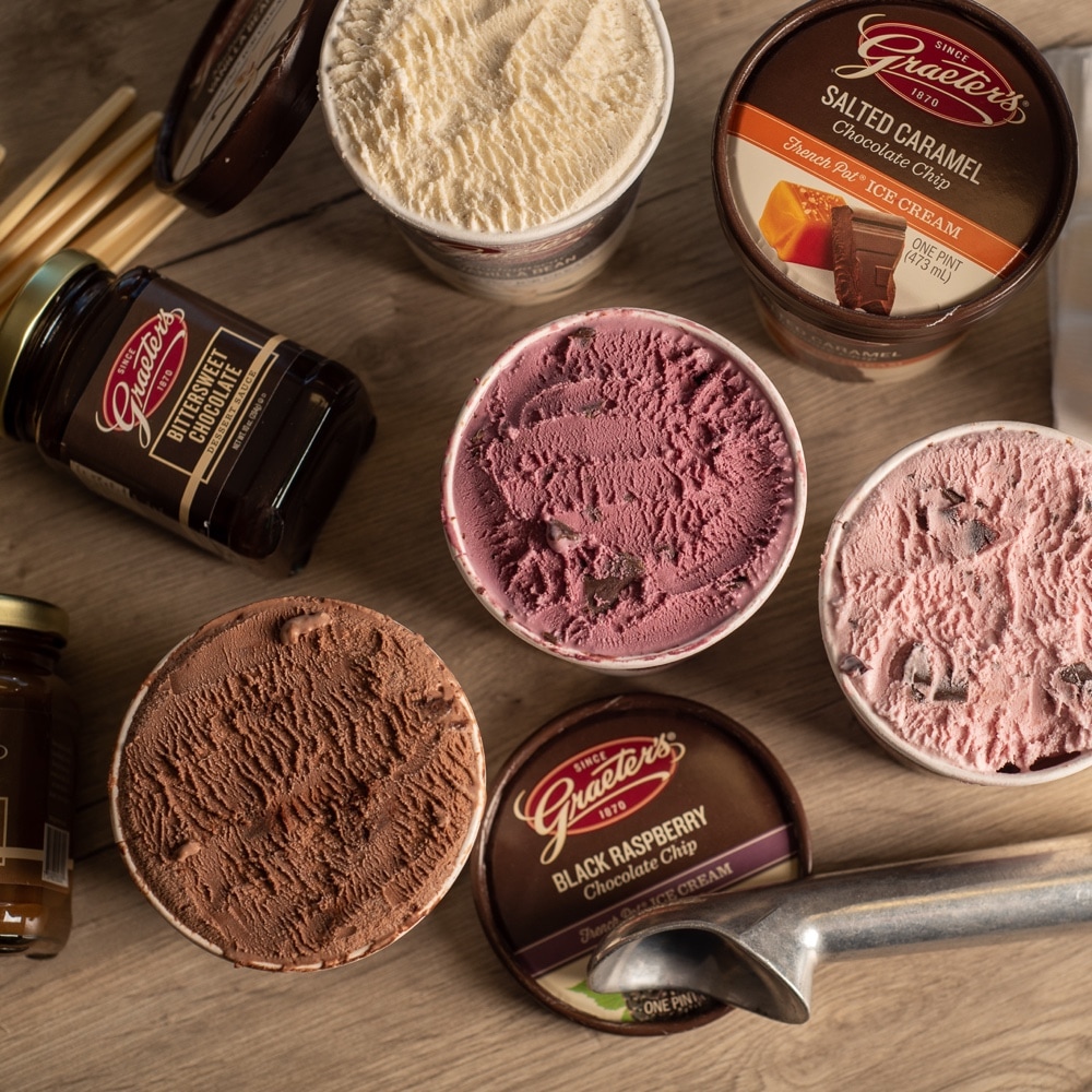Four different pints of Graeter's Ice cream open with a metal ice cream scoop laid in front, Grater's Butterscotch Dessert Sauce & Bittersweet Dessert Sauce lay on their sides and a stack of disposable ice cream bowls that say "Grater's since 1870" lay on their side.