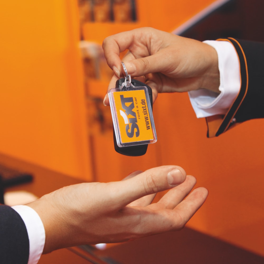 Eine SIXT-Mitarbeiterin legt einem Kund einen Schlüsselbund mit SIXT-Logo-Schlüsselanhänger in die wartenden Hände.