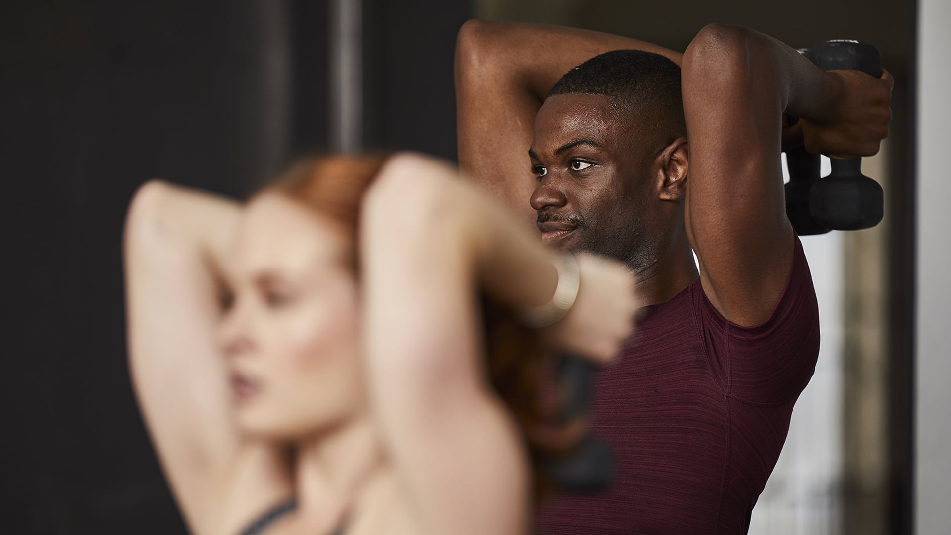 A woman holds a dumbbell behind her head while a man in the background does the same