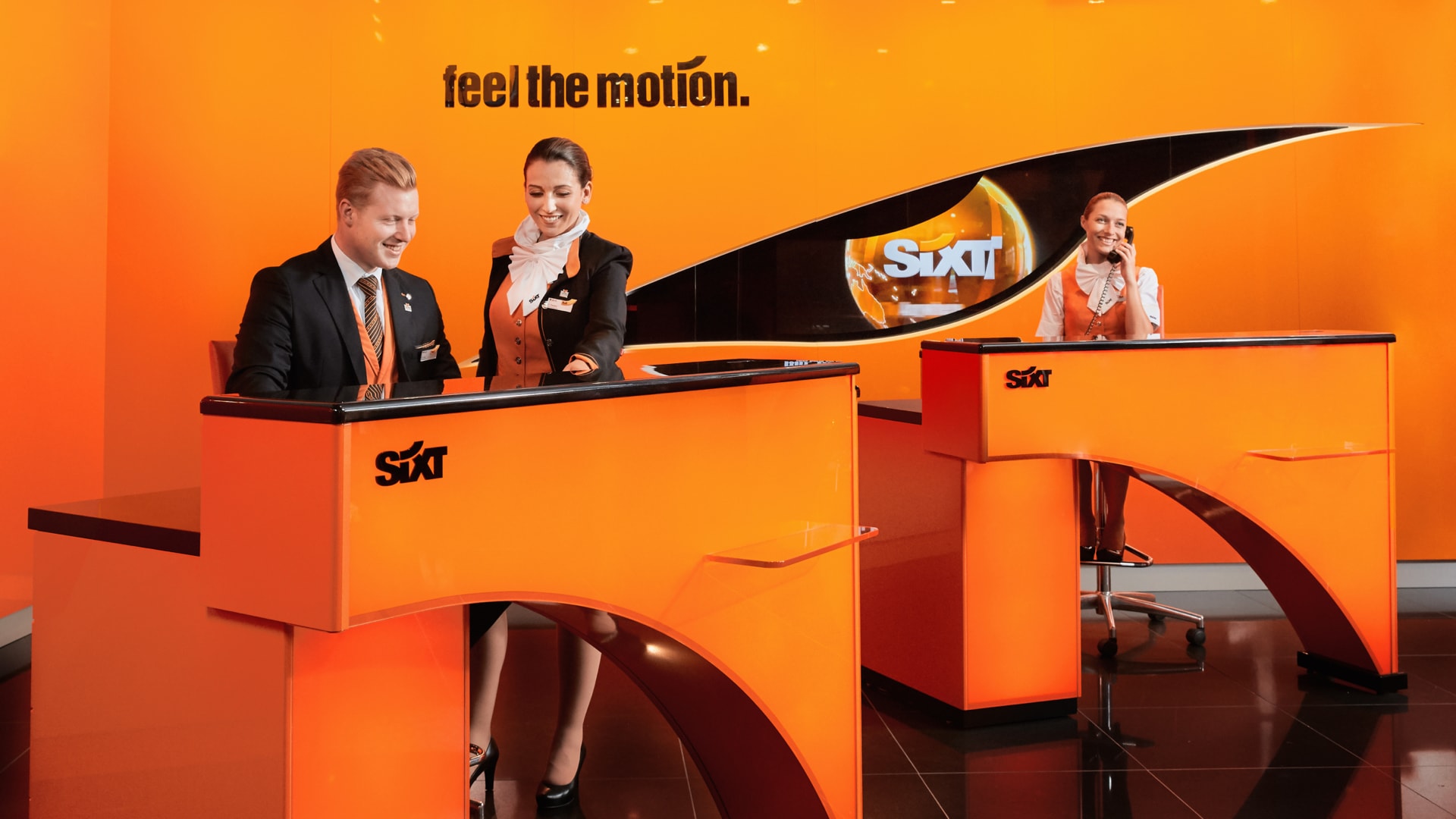 Drei SIXT-Mitarbeiter*innen in schwarzer und orangefarbener Geschäftskleidung arbeiten vor einer orangefarbenen Wand, auf der in schwarzer Schrift „feel the motion“ steht.